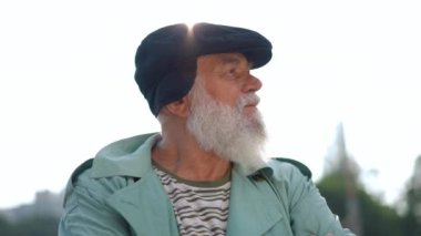 Thoughtful old man standing in sunbeam outdoors looking away turning looking up in slow motion. Close-up portrait of relaxed confident Caucasian bearded retiree at background of sunshine