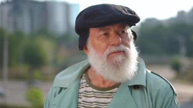 Close-up portrait of serious concentrated old bearded man looking around standing on city street at background of blurred town. Thoughtful Caucasian retiree outdoors on sunny day. Slow motion