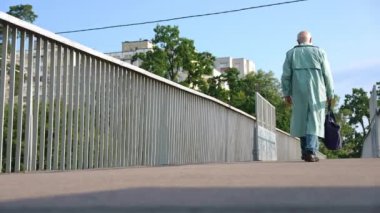 Wide shot old Caucasian gray-haired man with tote bag walking in slow motion on urban bridge on the right. Back view unrecognizable male retiree with groceries strolling in town outdoors