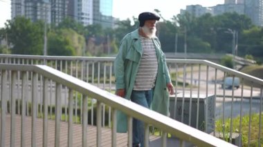 Live camera follows confident old man walking downstairs from pedestrian bridge strolling in urban city in slow motion. Side view tracking shot of Caucasian retiree outdoors in town
