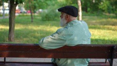 Back view medium shot of senior bearded man in hat and cloak sitting on bench turning looking around. Relaxed Caucasian retiree resting in sunny summer spring park outdoors thinking