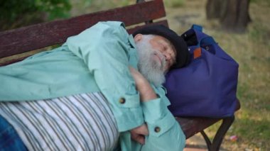 Portrait of senior sleeping homeless man waking up sitting down on bench in park looking away. Sad depressed helpless Caucasian old retiree outdoors. Social issues and inequality concept
