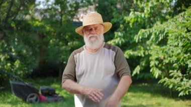 Confident senior gardener crossing hands looking at camera standing on sunny summer spring day outdoors. Medium shot front view portrait of handsome Caucasian old man posing outdoors