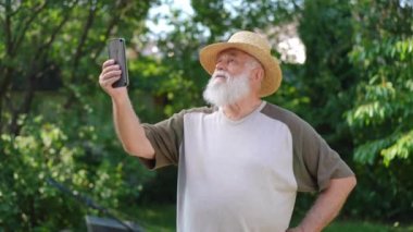 Hasır şapkalı yaşlı beyaz çiftçi dışarıda duran akıllı telefon uygulamasında görüntülü sohbet kullanarak konuşuyor. Çevrimiçi bahar bahçesinde son sınıf özgüvenli bahçıvan blogu portresi