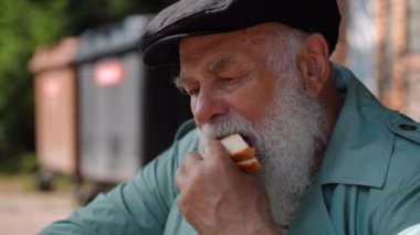 Close-up homeless old man enjoying tasty sandwich gesturing thumb up talking in slow motion. Portrait of lonely senior Caucasian retiree eating sitting outdoors on city street