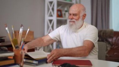 Positive senior bearded man sitting at table with books and laptop smiling. Confident Caucasian old male retiree enjoying leisure indoors at home in living room