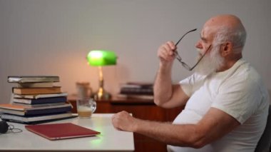 Side view of bearded senior man putting on eyeglasses counting books on table turning looking at camera. Serious intelligent Caucasian retiree posing on the right indoors at home
