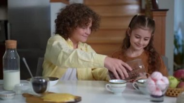Smiling brother pouring dry breakfast in plates for sister and himself sitting at table at home. Portrait of happy positive Caucasian boy and girl eating healthful cereals in the morning