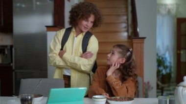 Confident brother with crossed hands asking sister to eat morning cereals before school. Positive loving Caucasian boy talking with girl at home indoors. Slow motion