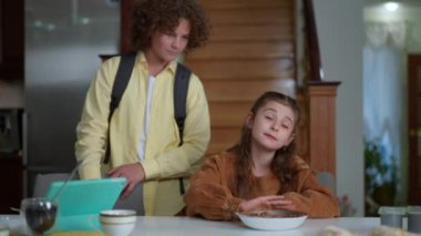 Beautiful Caucasian girl refusing eating breakfast cereal as smiling boy talking standing aside. Portrait of capricious sister sitting at table at home as brother waiting for going to school