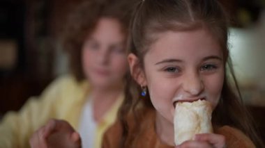 Close-up joyful brunette Caucasian girl looking at camera eating tasty bun laughing. Blurred boy smiling sitting at background. Portrait of relaxed happy sister dining with brother posing