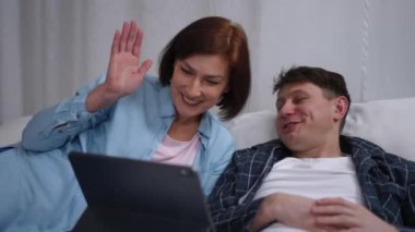 Portrait of smiling man in pajamas and woman in blazer waving talking using video chat app on digital tablet lying in bed. Ill Caucasian husband and caring wife messaging in web chat at home