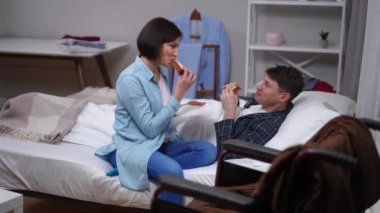 Wide shot positive husband and wife eating sandwiches talking smiling. Ill Caucasian man lying in bed chatting with loving woman in bedroom with wheelchair. Relationship and lifestyle