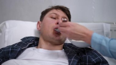 Portrait of fatigue sick man lying in bed as female hands feeding porridge with spoon wiping spilled milk on face. Thankful Caucasian husband looking at loving unrecognizable wife smiling