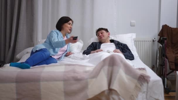 宽阔的前景快乐的妻子和生病的丈夫躺在床上 用遥控式的交谈切换电视频道 积极的白人女性和男性 除了坐轮椅 在卧室享受休闲 — 图库视频影像