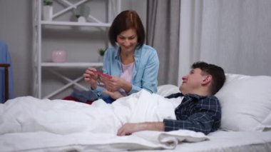Smiling Caucasian woman filing nails for ill man lying in bed talking. Portrait of caring wife enjoying leisure with sick husband in bedroom at home indoors