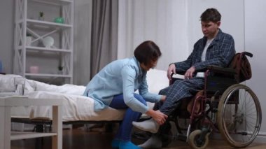 Tekerlekli sandalyedeki kocaya yardım eden, ayakları üzerinde duran, sevgi dolu bir eş, beşlik çakıyor. Beyaz bir kadın ve erkek evlerinde beraberler.