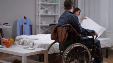 Pijamalı, tekerlekli sandalyede oturan, arka planda yatağını yapan bulanık kadını bekleyen beyaz bir adam. Evde felçli bir koca ve sevgi dolu bir eş.