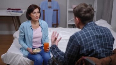 Rack focus from desperate caring wife persuading husband in wheelchair to eat porridge with juice to angry spouse yelling. Stressed crying woman leaving bedroom