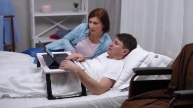 Smiling woman entering bedroom lying down to ill man with digital tablet typing on keyboard talking. Dissatisfied Caucasian irritated husband shaking head with disapproving facial expression