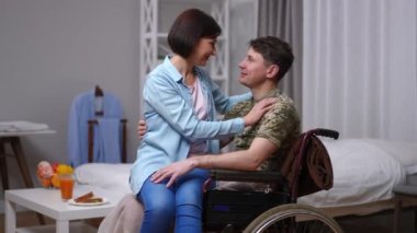 Smiling happy woman sitting on lap of man in wheelchair rubbing noses. Side view portrait of loving Caucasian husband and wife at home indoors enjoying leisure. Bonding and disability