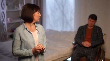 Depressed woman praying standing in bedroom with blurred desperate man in wheelchair at background. Portrait of stressed worried Caucasian wife asking God for help for sick husband