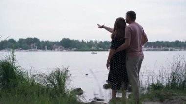 Wide shot young Caucasian couple standing on the right on river bank pointing away waving. Happy loving man and woman hugging enjoying dating outdoors on spring summer day. Relationship concept