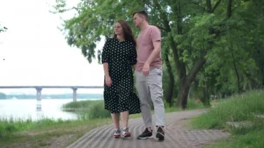 Wide shot happy hugging couple walking on park alley talking looking away pointing. Dolly shot portrait of loving Caucasian boyfriend and girlfriend strolling dating outdoors on spring summer day
