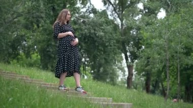 Wide shot beautiful young pregnant woman walking downstairs in spring summer park. Portrait of charming confident Caucasian expectant enjoying leisure outdoors. Maternity and nature concept