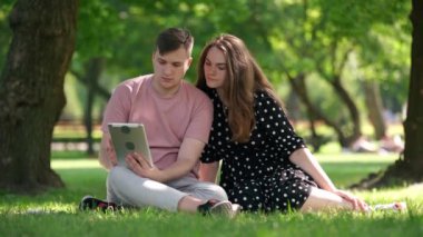 Absorbed gen Z couple scrolling social media in digital tablet app talking sitting on sunny meadow in summer spring park. Wide shot young Caucasian man and woman talking enjoying weekend leisure