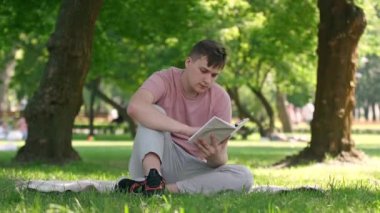 Portrait of concentrated absorbed young man reading book turning page sitting on summer spring meadow in park. Intelligent Caucasian reader enjoying hobby outdoors on weekend