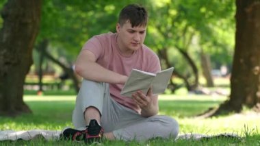 Young handsome man reading book looking around in sunny summer spring park. Portrait of absorbed happy relaxed guy enjoying hobby on weekend outdoors