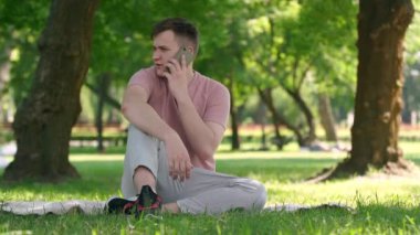 Cheerful relaxed young man picking up phone talking laughing sitting in sunny park outdoors. Wide shot portrait of confident Caucasian guy chatting enjoying weekend leisure on summer spring day