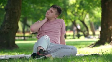 Portrait of young Caucasian man stretching painful neck sitting on sunny lawn in spring summer park. Wide shot front view of gen Z guy resting outdoors on weekend. Life balance concept