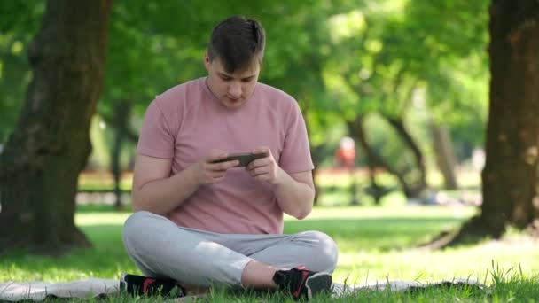 スマートフォンアプリでワイドショット集中ゲンZ男ゲームは手に頭を持って携帯電話の観光を保持置くを失う 日当たりの良い夏の公園で不満若いゲーマーの屋外の肖像画 — ストック動画