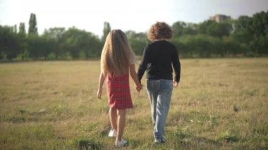 Wide shot carefree happy children walking on sunny spring summer meadow holding hands. Back view relaxed Caucasian boy and girl strolling outdoors at sunset enjoying weekend