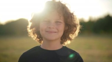 Close-up portrait of confident happy Caucasian boy with curly hair in sunrays smiling looking at camera. Carefree relaxed child posing in sunshine outdoors at sunset