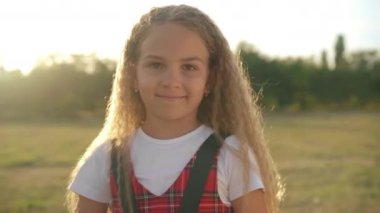 Portrait of carefree charming Caucasian girl in sunrays laughing looking at camera. Happy confident kid posing in sunset sunshine outdoors on meadow enjoying weekend leisure on spring summer day