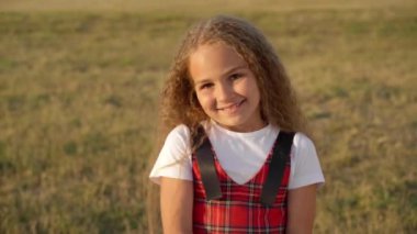 Beautiful Caucasian girl smiling looking at camera standing in sunshine on spring summer meadow. Portrait of confident cute kid posing at sunset outdoors. Childhood and individuality concept