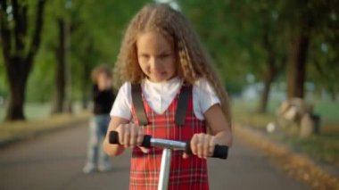 Portrait of excited charming girl riding kick scooter on park alley with blurred boy clapping at background. Confident curious Caucasian kid learning to ride on summer spring day outdoors
