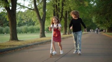 Happy smiling Caucasian girl riding kick scooter with boy running aside leaving with friend. Excited friends having fun on spring summer day on park alley outdoors