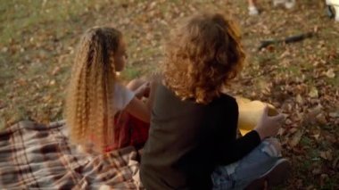 High angle view carefree Caucasian boy and girl sitting on blanket on autumn meadow with ukulele. Wide shot happy relaxed brother and sister enjoying leisure on weekend outdoors