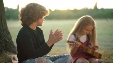 Side view positive boy clapping as blurred girl learning playing ukulele at sunset outdoors. Caucasian kid endorsing supporting friend sitting on meadow with string instrument. Hobby and talent