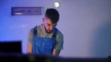 Tattooed young man entering garage opening car hood leaning to engine. Portrait of Caucasian mechanic repairing vehicle breakdown. Service and maintenance concept