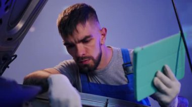 Troubled auto mechanic with digital tablet holding head in hands examining car engine. Portrait of young Caucasian man thinking fixing automobile breakdown at service station