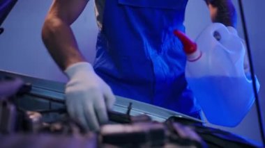 Unrecognizable man pouring antifreeze coolant in car hood indoors. Front view Caucasian young tattooed auto mechanic doing maintenance for vehicle at service station