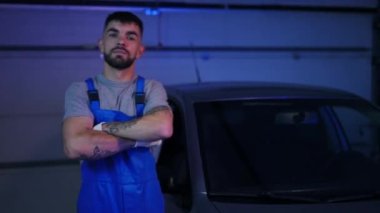 Confident positive young man crossing hands looking at camera smiling standing in garage at car on the left. Portrait of professional Caucasian auto mechanic posing at service station indoors