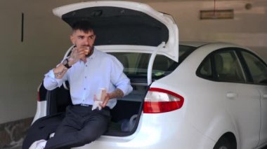 Tattooed young man eating sandwich sitting on car trunk in garage. Portrait of relaxed Caucasian guy enjoying snack indoors