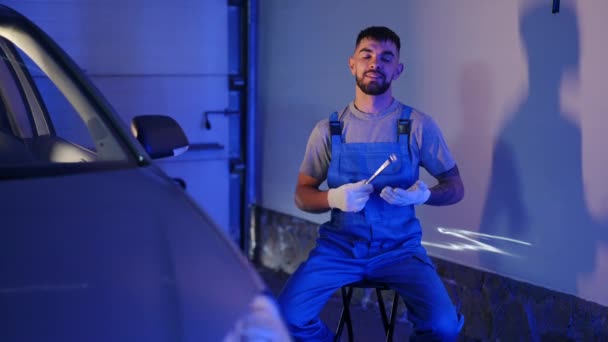 Portrait Bearded Confident Young Man Uniform Wrench Sitting Garage Looking — Vídeo de stock