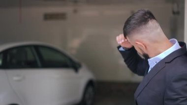 Thoughtful sad man standing on the right with car with flashing hazard lights at background. Stressed troubled Caucasian guy waiting for insurance agent in garage indoors looking away